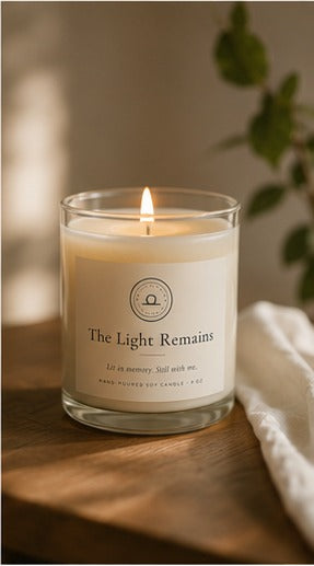 The Light Remains memorial soy candle burning on a wooden table beside a green plant