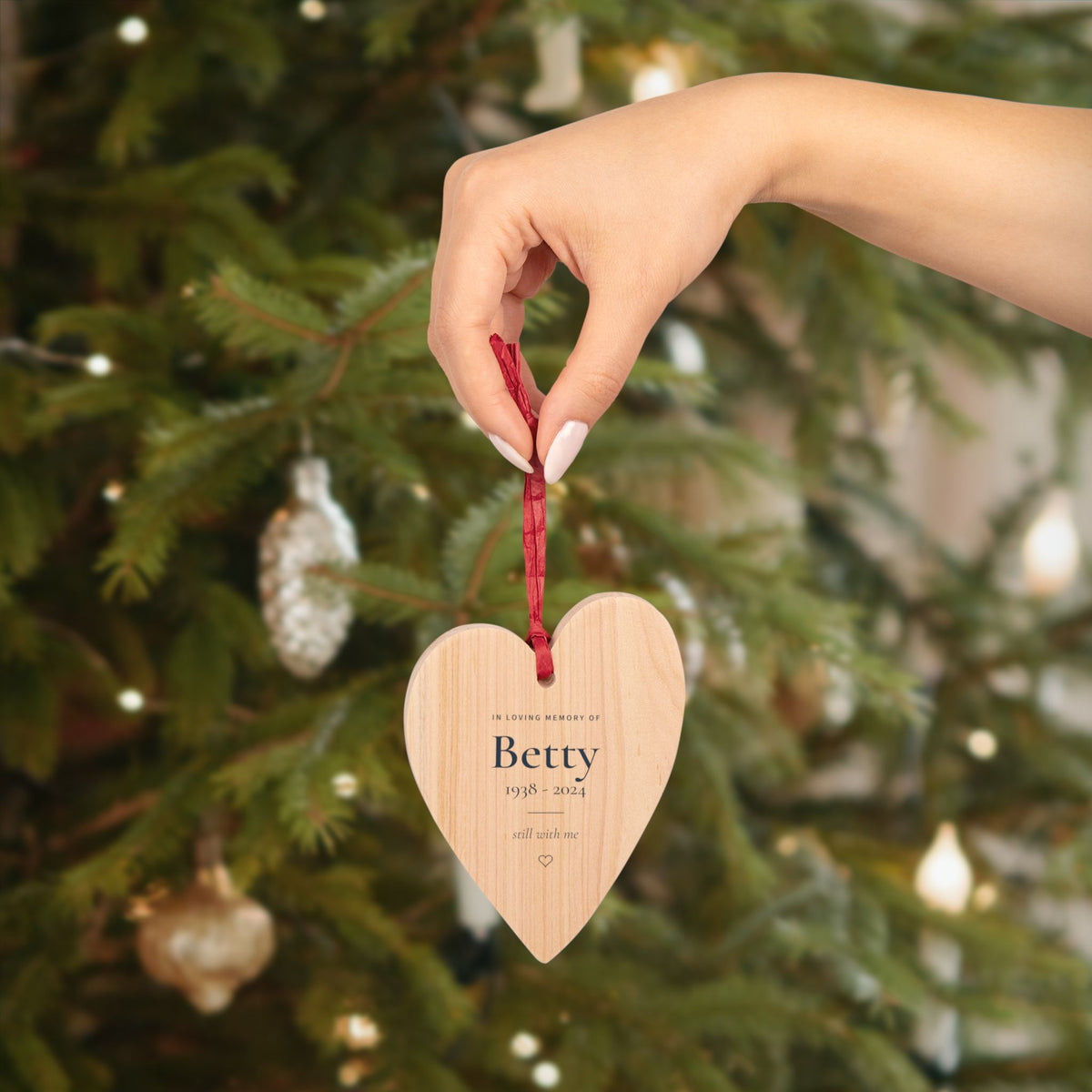Hand holding the personalized wooden heart memorial ornament with the name Betty in front of a Christmas tree